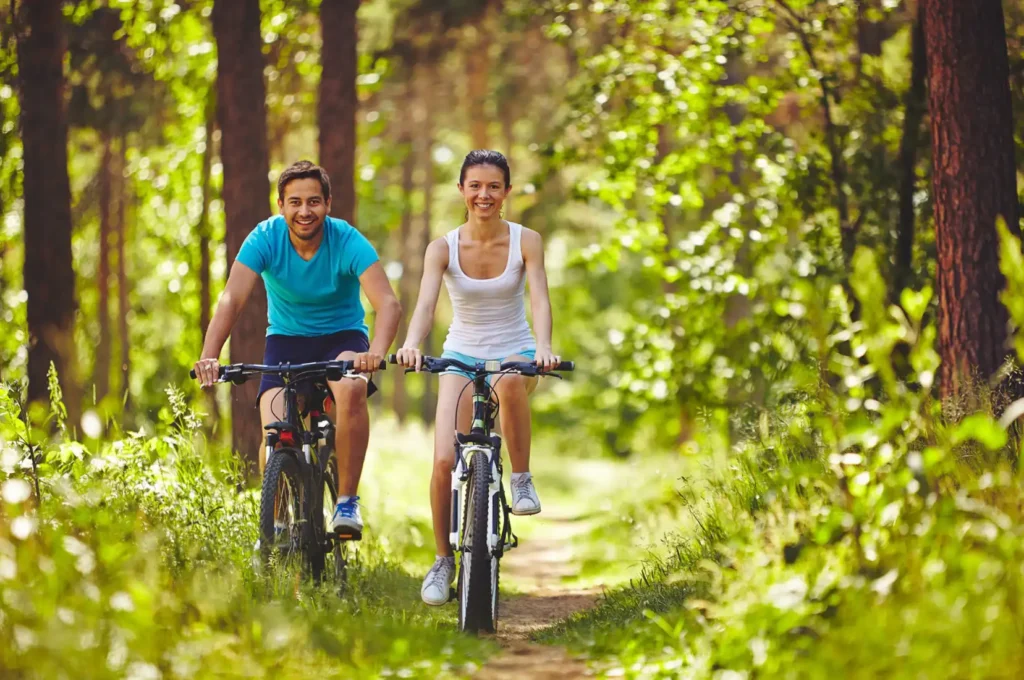 A woman and a man wearing workout clothes biking on a trail through the woods.