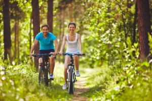 A woman and a man wearing workout clothes biking on a trail through the woods.
